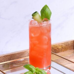 Tall glass of colorful cocktail with basil and cucumber garnish on reflective tray, perfect for summer refreshment.